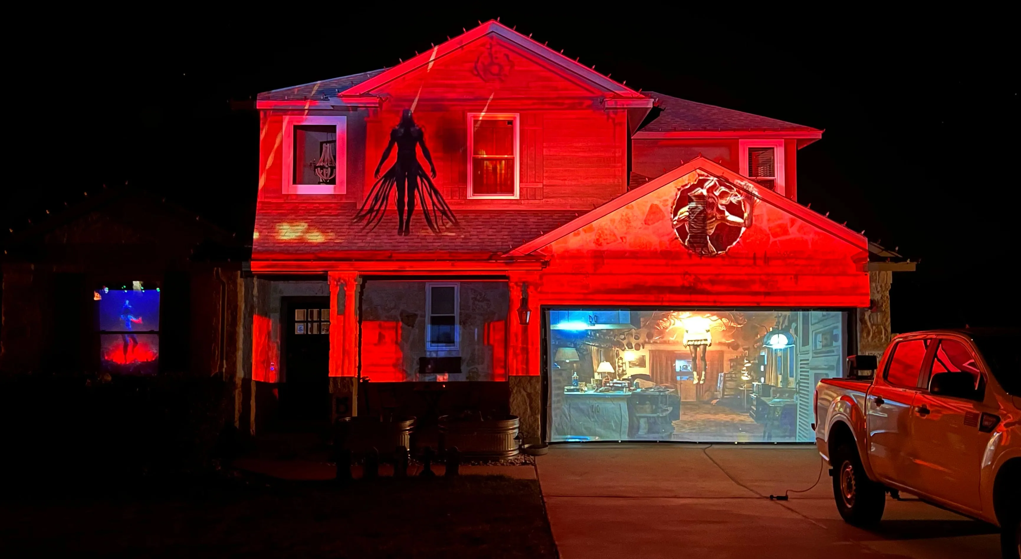 A two-story house at night washed in red light, with a tall shadowy figure projected across the upper story and a lit living-room scene projected onto a screen over the garage door.