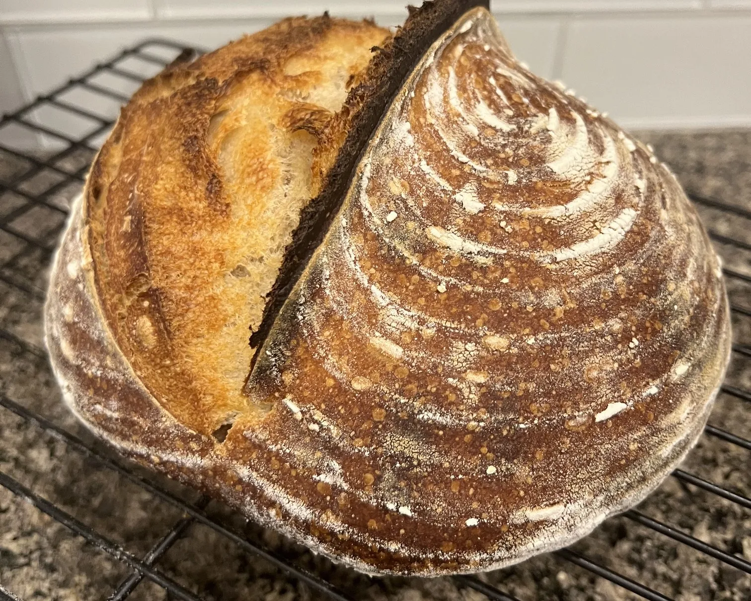 A round sourdough loaf on a cooling rack, with flour-dusted spiral rings from the banneton and a single deep score that has opened into a raised ear.