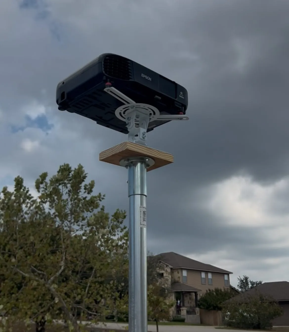 An Epson projector on a low-profile mount, bolted through a wooden plate to a floor flange on top of a galvanized fence post, against a cloudy sky.
